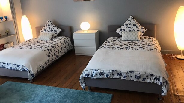 A bedroom with two beds, each with a white and blue floral bedspread, flanked by bedside tables with lamps. A small painting hangs on the wall above the beds. - EGLANTINE  (Dinan)