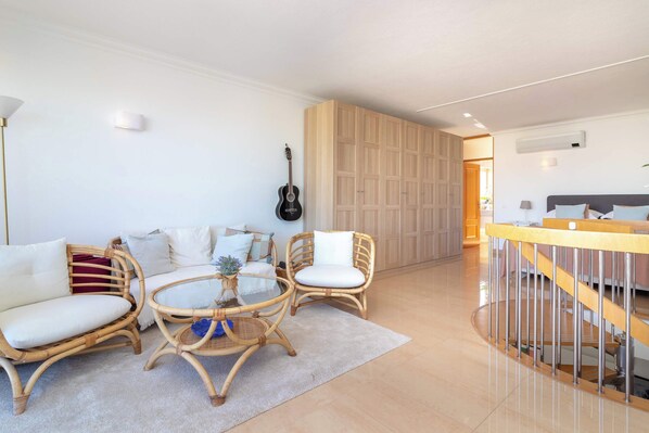 A modern living room with a sofa, two wicker chairs, a glass coffee table, and a guitar on the wall.
