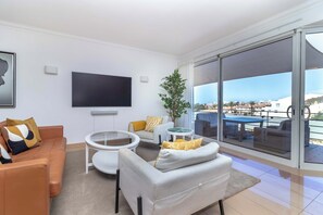 A modern living room with a flat-screen TV, a sofa, a coffee table, and a view of the city through large windows. - Parque nº 8 - Two Bedroom Penthouse Apartment (Ferragudo)