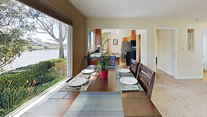 A dining area with a view of a lake, a kitchen in the background, and a carpeted floor. - Private Beach House with amazing views of Shwan Lake and Twin Lakes Beach (Santa Cruz)
