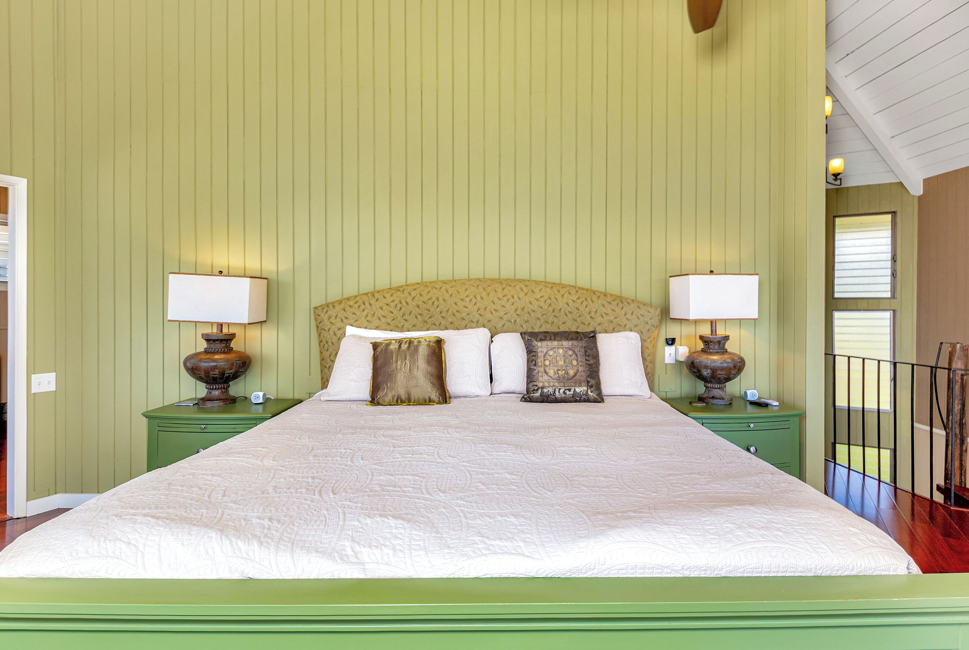 Een net opgemaakt bed met een hoofdbord, geflankeerd door twee nachtkastjes met lampen, in een kamer met groene wanden en een houten vloer.