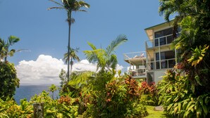 A modern two-story building with a balcony, surrounded by lush tropical vegetation and a view of the ocean. - Paradise Bluff: Cliffside Home w/Gorgeous Ocean Views and Tropical Garden (Laupahoehoe)