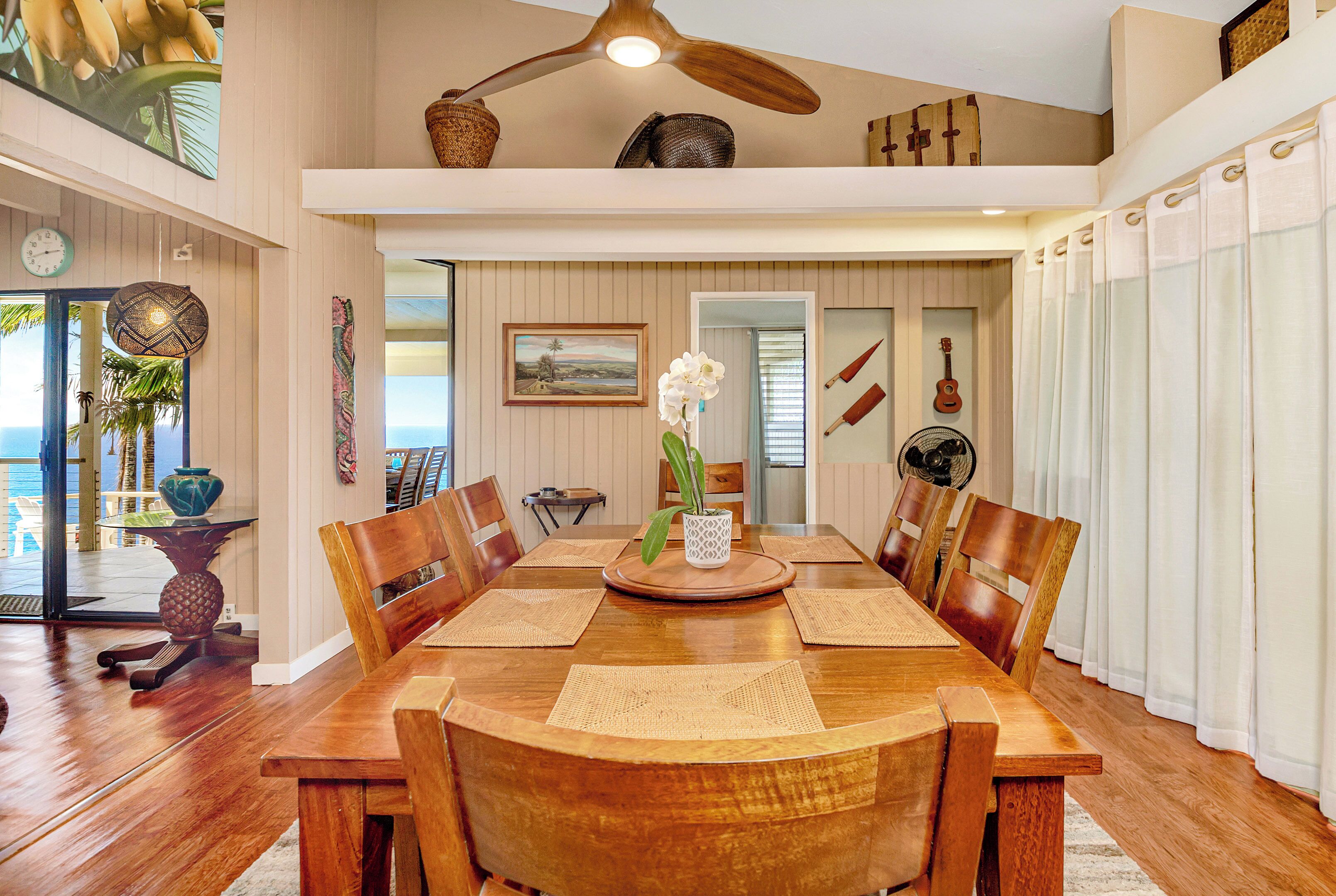 A spacious dining area with a wooden table and chairs, a ceiling fan, and a view of the ocean through large windows.