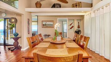 A spacious dining area with a wooden table and chairs, a ceiling fan, and a view of the ocean through large windows.