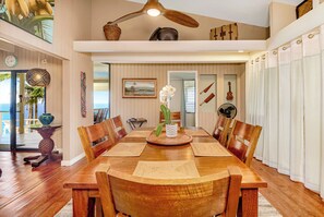 Uma sala de jantar ampla com uma mesa e cadeiras de madeira, um ventilador de teto e vista para o mar através de grandes janelas.