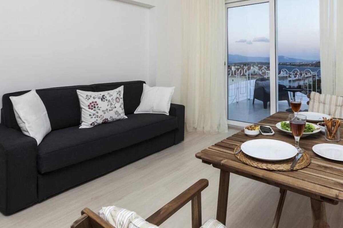 A modern living room with a black sofa, white cushions, a wooden dining table with plates and cutlery, and a large sliding glass door leading to a balcony with a view.