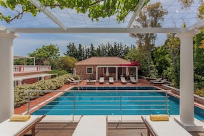 En swimmingpool med solstolar och en pergola, omgiven av grönska och en byggnad i tegel.