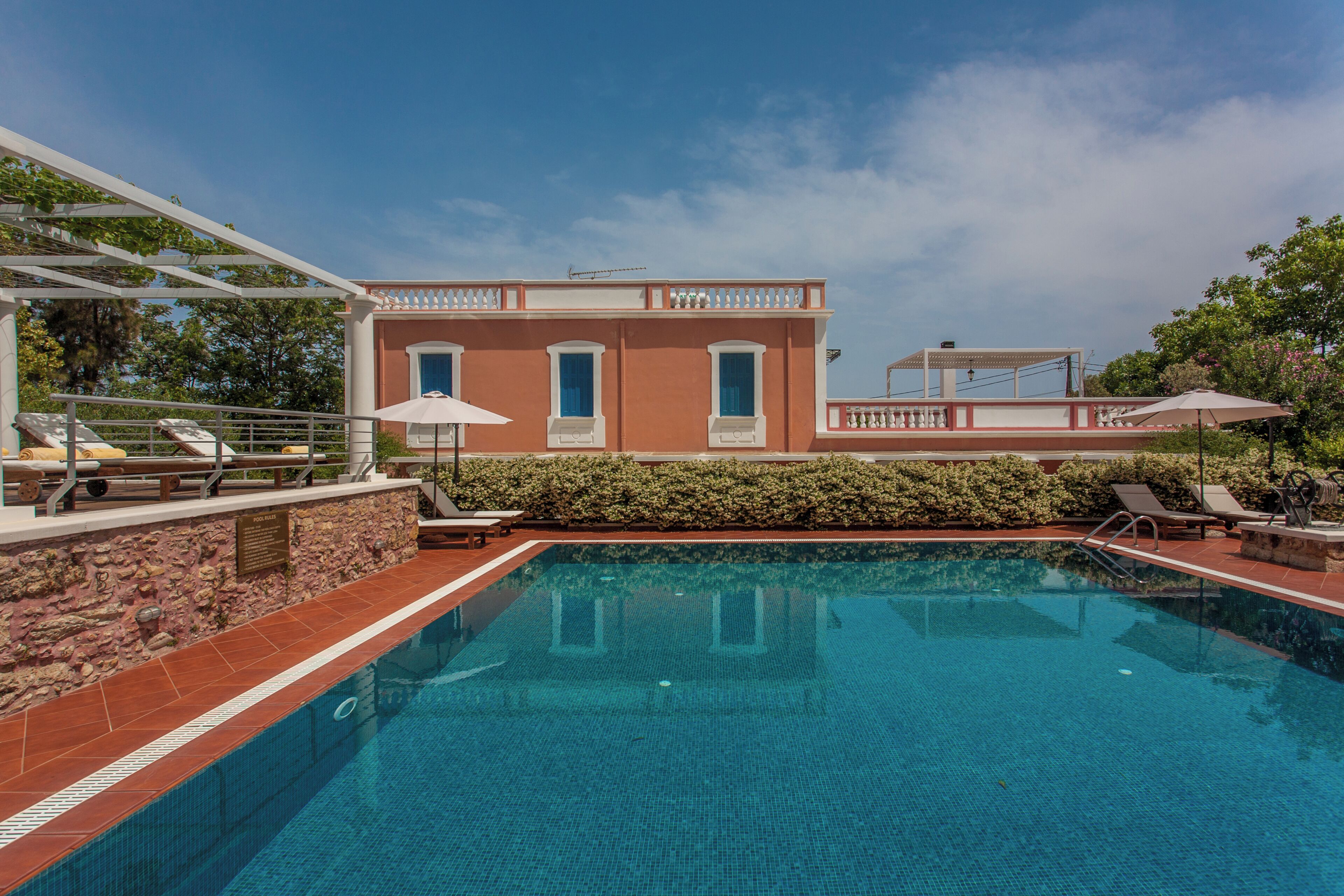 Une piscine bordée d’un mur en pierre, un bâtiment doté d’un balcon et un ciel d’un bleu limpide.