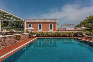 A swimming pool with a stone wall, a building with a balcony, and a clear blue sky.