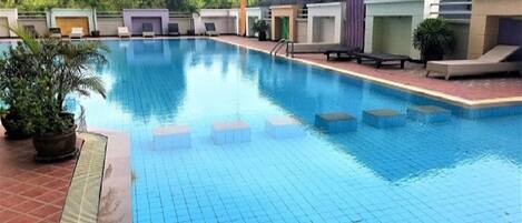 A swimming pool with tiled flooring, lounge chairs, and a building with a flat roof in the background.