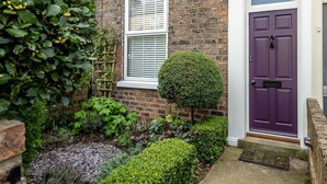 A brick house with a purple door, a window with shutters, and a well-maintained garden with various plants and a neatly trimmed hedge. - 3 Darnborough Street -  a family break that sleeps 4 guests  in 2 bedrooms (York)