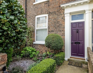 A brick house with a purple door, a white window, and a well-maintained garden with various plants. - 3 Darnborough Street - a family break that sleeps 4 guests in 2 bedrooms (York)