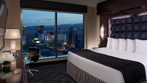 A hotel room with a large bed, a city view, a desk with a lamp, and a chair.