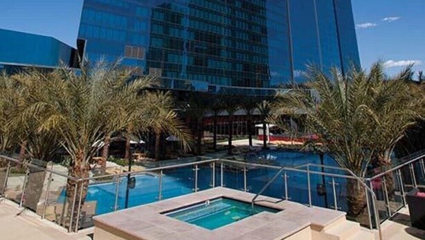 A modern building with a reflective glass facade, a pool area with palm trees, and a clear blue sky.