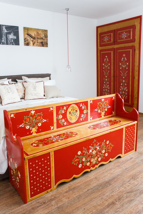 Une pièce comprenant un lit, un banc rouge orné de motifs floraux, une armoire en bois et deux tableaux encadrés accrochés au mur.
