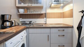 Cocina compacta con lavadora integrada, cafetera y repisa de vidrio con utensilios de cocina.