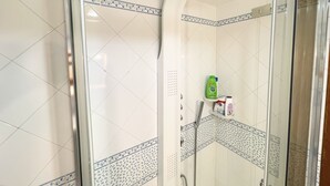 A shower with a glass door, white tiles with blue floral borders, and a shelf with toiletries.