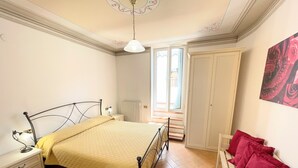 A bedroom with a bed, bedside table, nightstand, radiator, window, and a chair with red cushions.