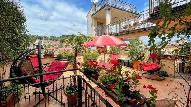 A rooftop terrace with red chairs, a red umbrella, potted plants, and a view of the cityscape.