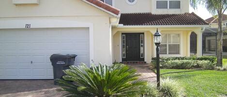Una casa de dos pisos con garaje, puerta principal y un pequeño jardín con una palmera.