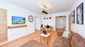 A modern living room with a wooden floor, a brown sofa, a TV, a clock, and a dining area with chairs and a table.