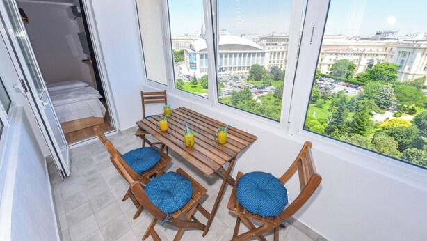 A room with a wooden table and four chairs, a bed, and a large window with a city view.
