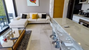 Ein modernes Wohnzimmer mit einer Couch, einem Glastisch und einer Küchenzeile, inklusive Kühlschrank und Mikrowelle.