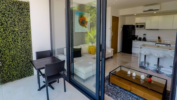 Ein modernes Wohnzimmer mit Glastür zum Balkon, ein Essbereich mit Tisch und Stühlen sowie eine Küche mit weißen Schränken und einem schwarzen Kühlschrank.