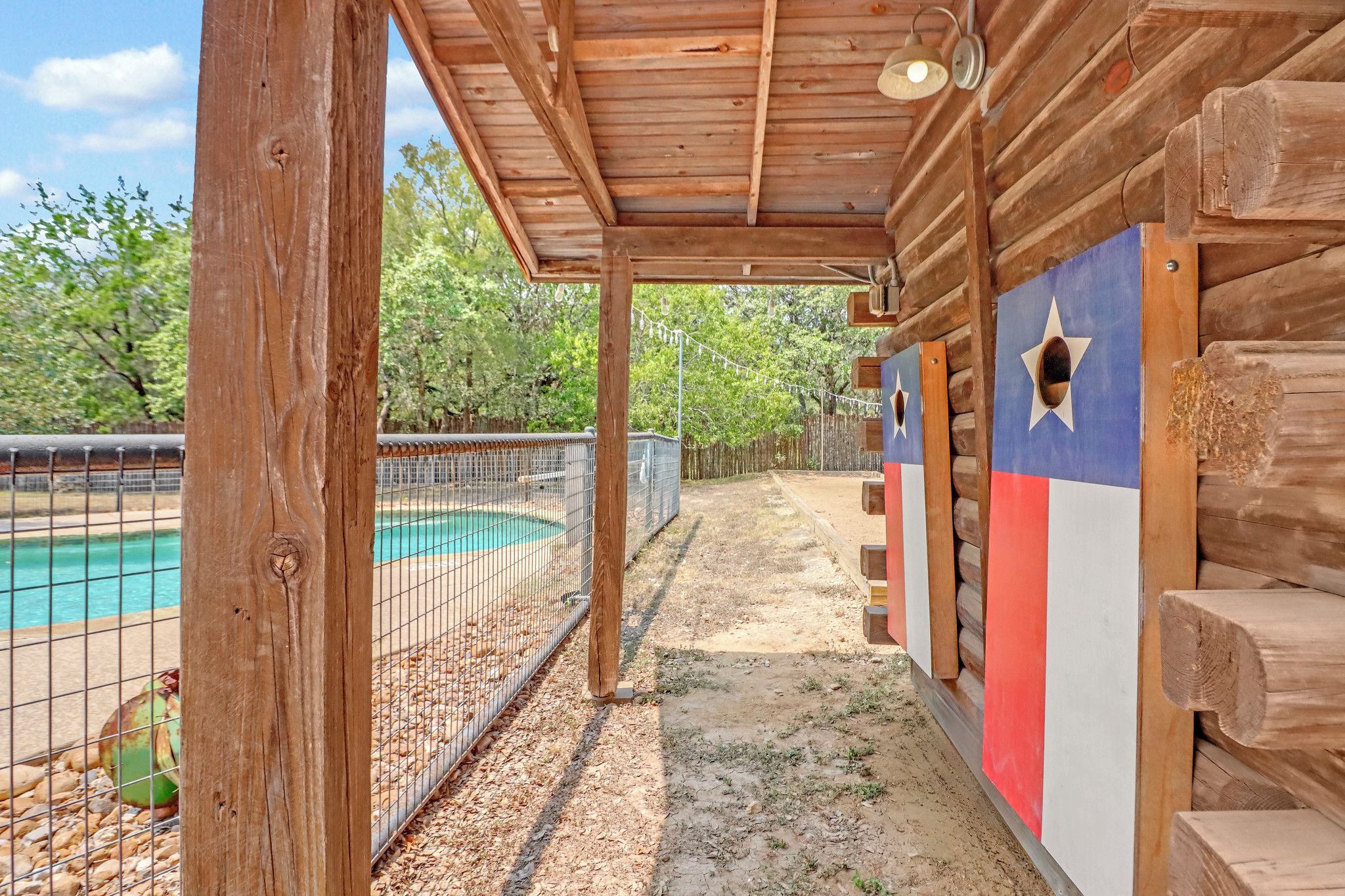 Una cabaña de madera con porche cubierto, piscina y un área cercada con árboles al fondo.