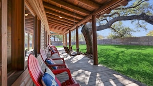 A covered outdoor seating area with wooden benches and a swing chair, surrounded by a wooden deck and a grassy lawn. - Resort Suite Across the Street from Market Days - Sleeps 4! (Wimberley)