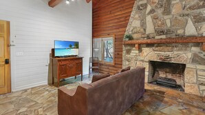 A cozy living room with a stone fireplace, wooden beams, a television, and a stone floor. - Resort Suite Across the Street from Market Days - Sleeps 4! (Wimberley)