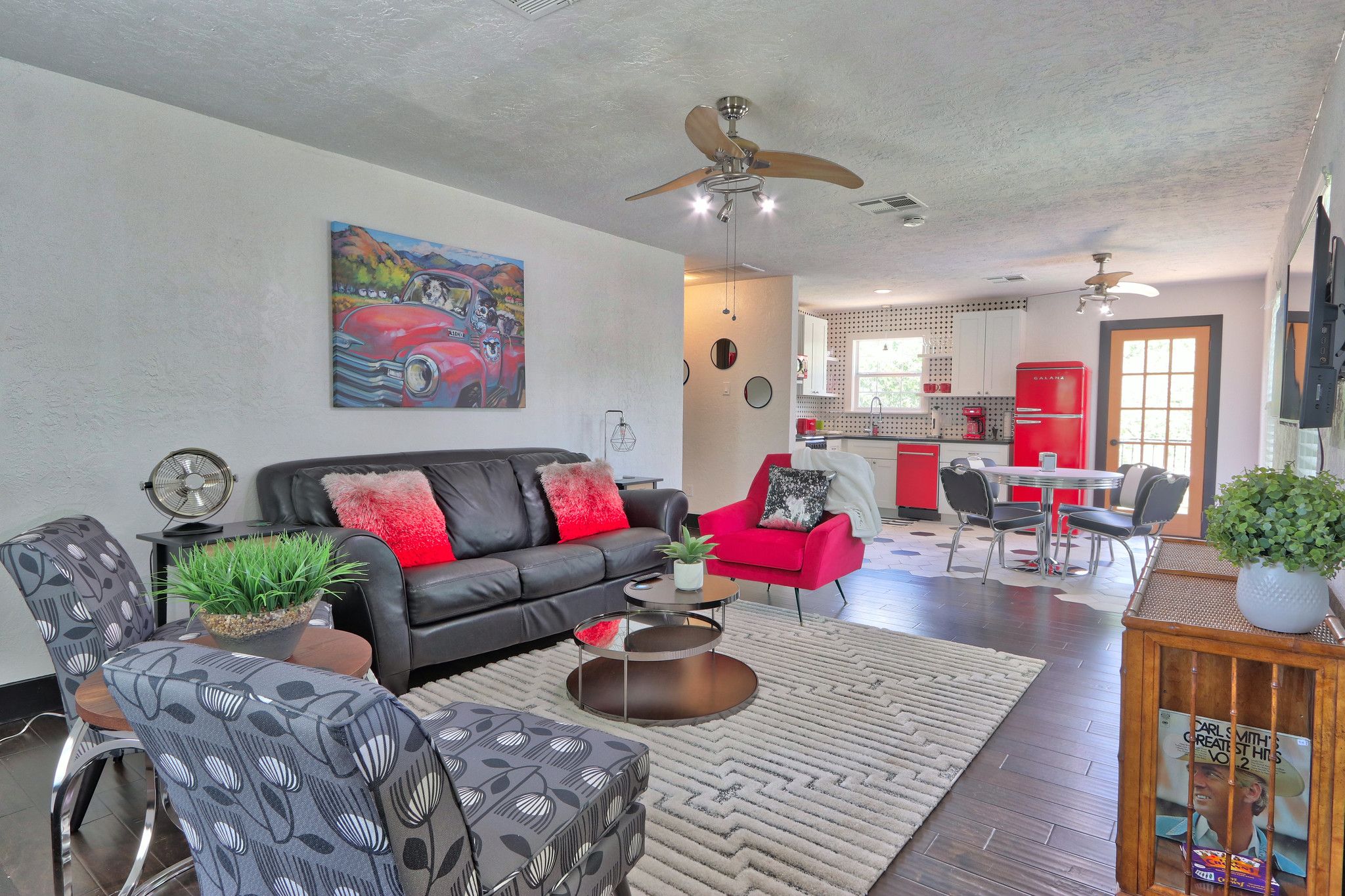 A modern living room with a black leather sofa, red armchair, and patterned chairs. A dining area with a red refrigerator and a kitchen in the background.