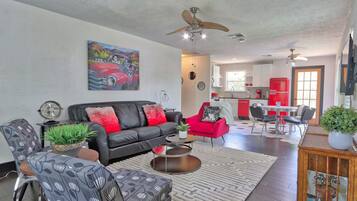A modern living room with a black leather sofa, red armchair, and patterned chairs. A dining area with a red refrigerator and a kitchen in the background.