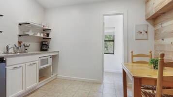 A kitchen with white cabinets, a microwave, and a coffee machine. There is a wooden dining table with chairs and a small plant. A door leads to a room with a window.