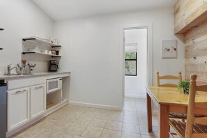 A kitchen with white cabinets, a microwave, and a coffee machine. There is a wooden dining table with chairs and a small plant. A door leads to a room with a window.