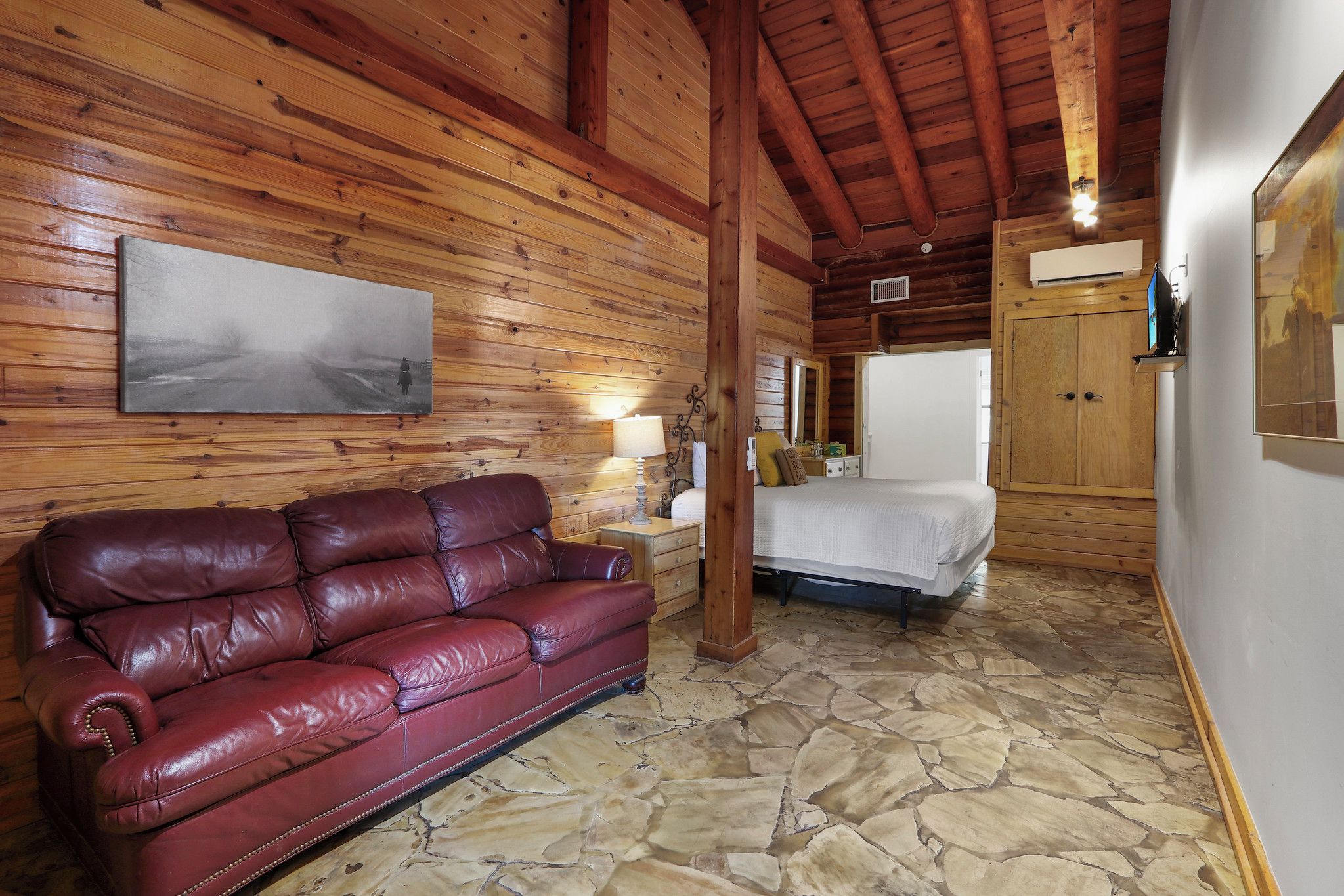A room with a stone floor, wooden walls, a red leather sofa, a bed, a nightstand with a lamp, and a painting on the wall.