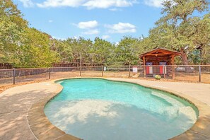 A clear swimming pool with a wooden structure nearby, surrounded by a fence and trees. - King Bed with Resort Pool and Firepit! Minutes from Downtown (Wimberley)