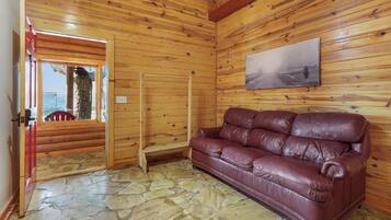 Interior de una cabaña de madera con piso de piedra, un sofá de cuero, una escalera de madera y un cuadro en la pared.