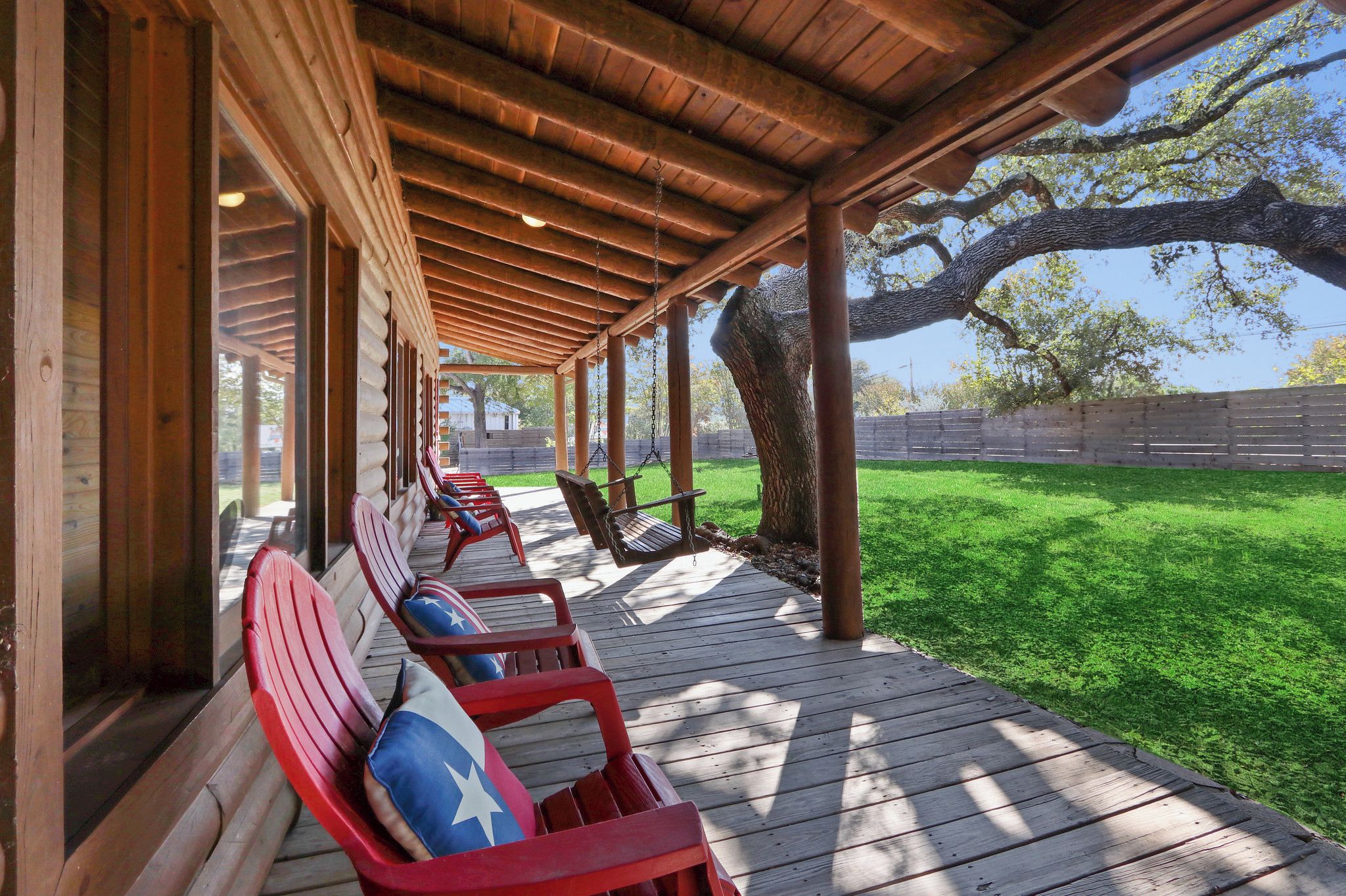 A covered outdoor seating area with wooden benches and a swing chair, surrounded by a wooden deck and a grassy lawn.