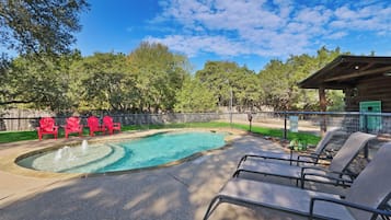 A swimming pool with a fountain, surrounded by red chairs and lounge chairs, with a wooden cabin in the background.
