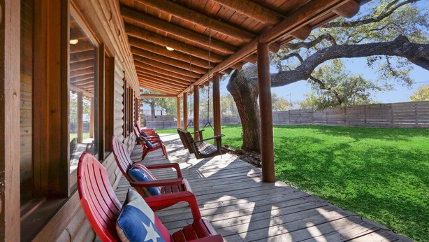 A covered outdoor seating area with wooden benches and a swing chair, surrounded by a wooden deck and a grassy lawn. - King Bed with Resort Pool and Firepit! Minutes from Downtown (Wimberley)