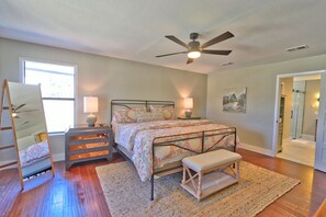 Un dormitorio con cama, lámparas de noche, cómoda de madera, banco, ventilador de techo, ventana y puerta que da a un baño.