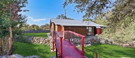 A wooden cabin with a red bridge, surrounded by a grassy area and trees.