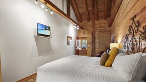 A bedroom with a large bed, wooden ceiling beams, a mounted TV, and a wooden wall with a decorative metal piece. - King Bed with Resort Pool and Firepit! Minutes from Downtown (Wimberley)