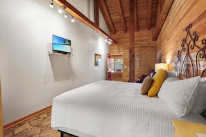 A bedroom with a large bed, wooden ceiling beams, a mounted TV, and a wooden wall with a decorative metal piece. - King Bed with Resort Pool and Firepit! Minutes from Downtown (Wimberley)