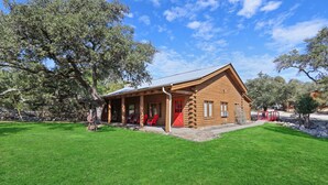 A log cabin with a porch and red door, surrounded by a grassy area and trees. - Hotel Style Suite with Resort Pool, Fire Pit, and Grill (Wimberley)