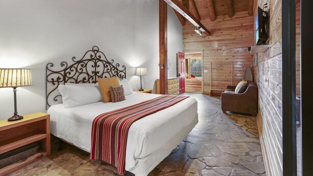A cozy bedroom with a wooden ceiling, a bed with a striped blanket, a nightstand with a lamp, and a wooden bench. - Hotel Style Suite with Resort Pool, Fire Pit, and Grill (Wimberley)