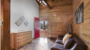 Un intérieur en bois avec un sol en pierre, une porte rouge, un canapé en cuir marron et une commode en bois.