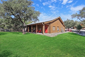 A log cabin with a porch and red door, surrounded by a grassy area and trees. - WLCRS - Unit 4 -Walk to Market Days -  Resort Pool! (Wimberley)