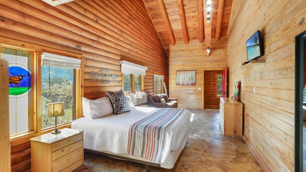 A log cabin bedroom with a large bed, wooden walls, and a stone floor. - WLCRS - Unit 4 -Walk to Market Days -  Resort Pool! (Wimberley)